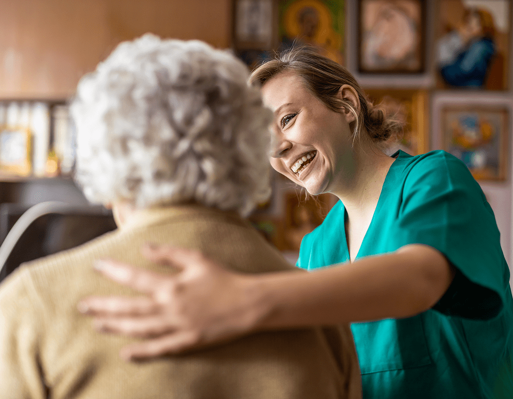 A young female caregiver in teal scrubs smiling warmly and embracing an elderly woman with white curly hair, in a care center setting with artwork on the wall in the background.