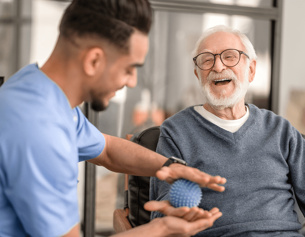 A healthcare worker in blue scrubs is using a textured blue ball to perform a hand exercise with an elderly man who is smiling and wearing glasses and a gray sweater.