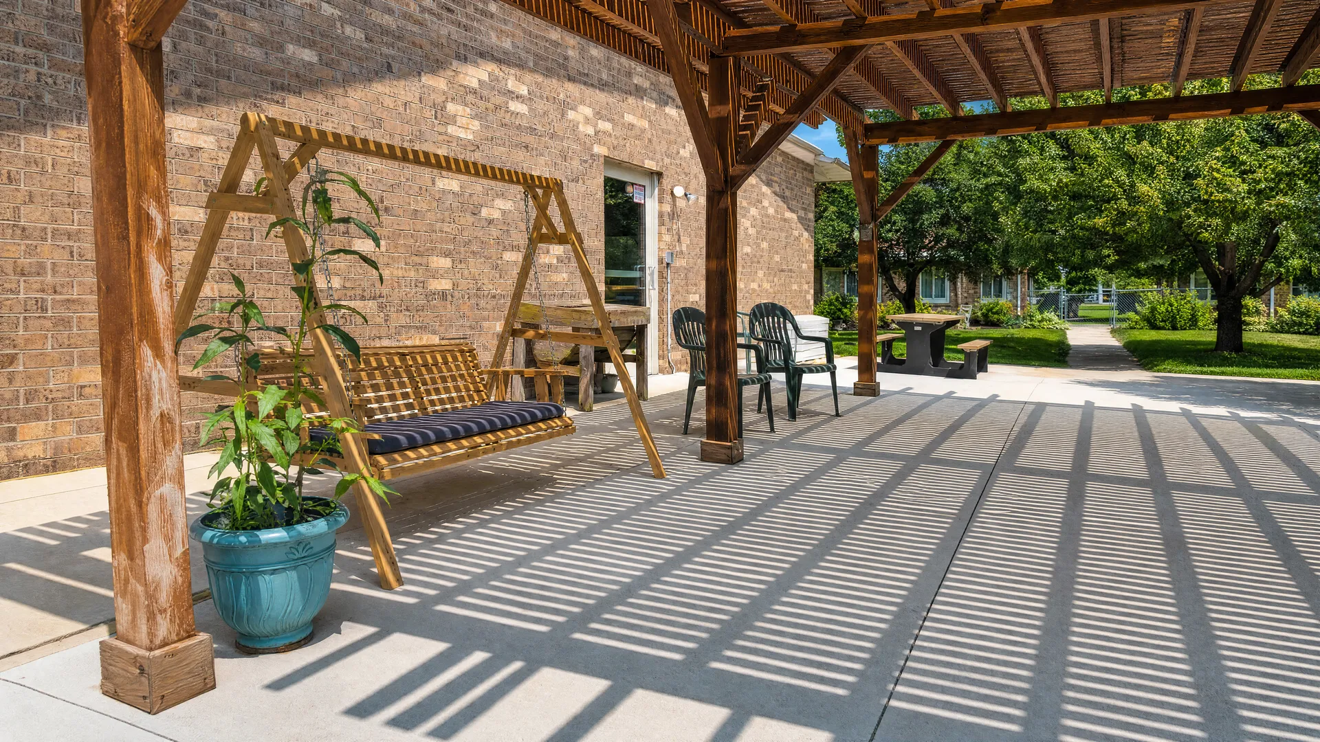 Wooden pergola patio with a porch swing and potted plants at Nora Springs Care Center